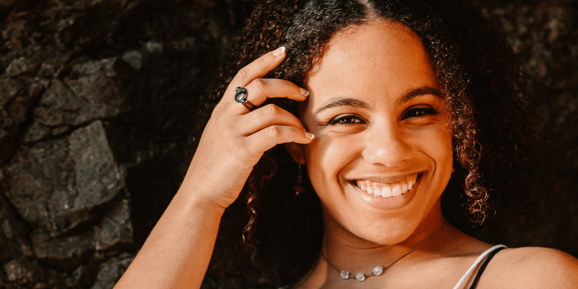 young woman with curly hair smiling at the camera in front of a rock wall. She's brushing her hair back from her face with a hand wearing a crystal conquering