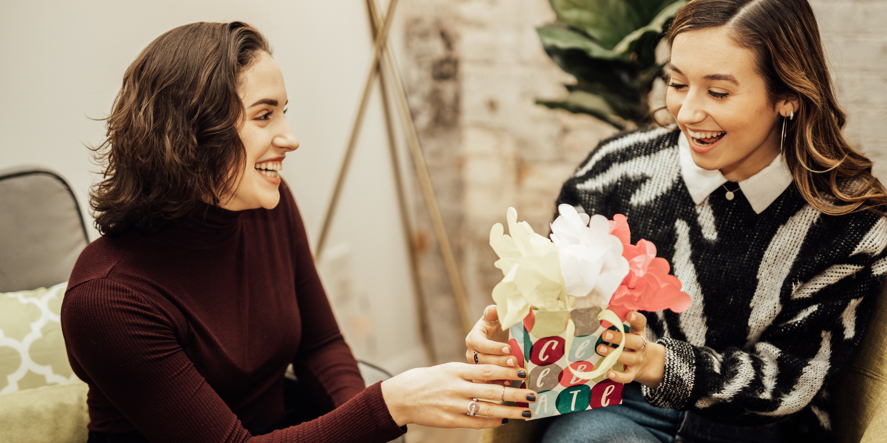 two young women sitting in chairs, one handing the other a colorful gift bag and both smiling