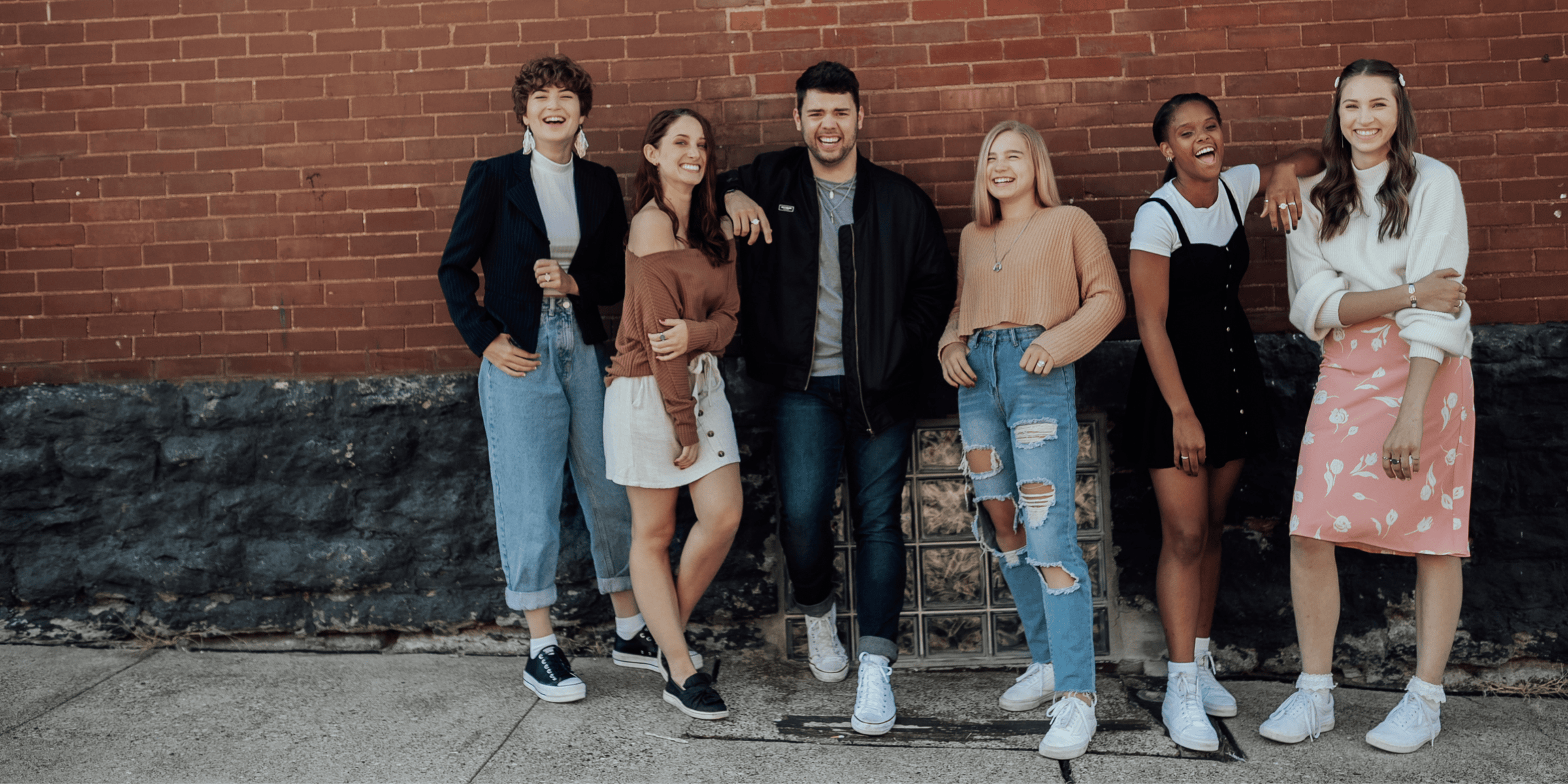 group of young women and one young man standing by a brick wall, smiling at the camera