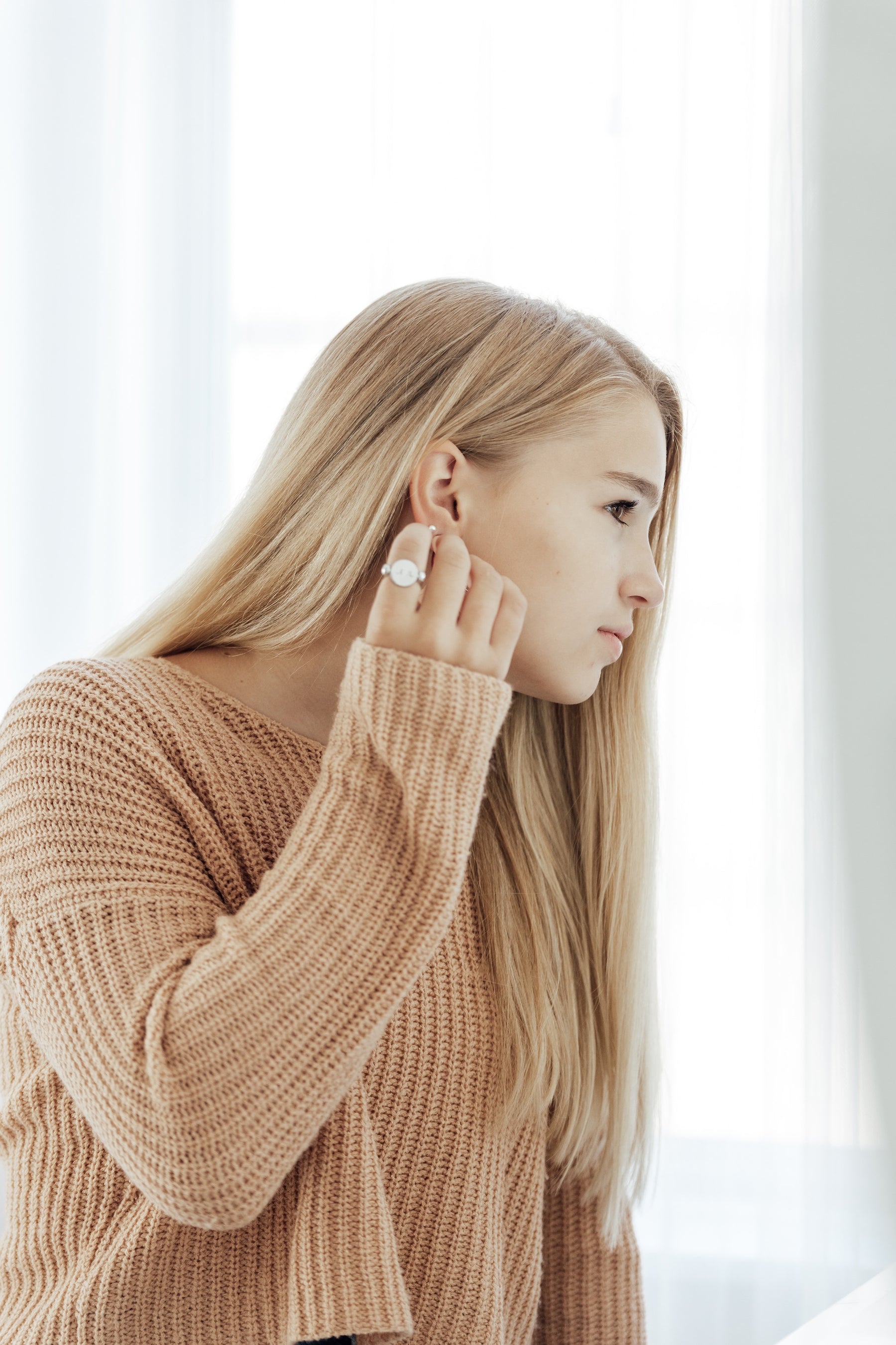 blond young woman adjusting earring while standing in front of a mirror, wearing a tan sweater and a silver conquering